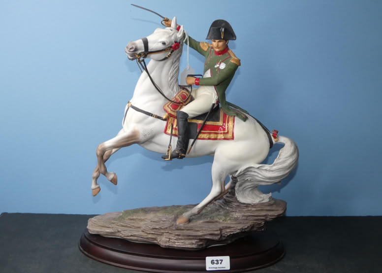 A figurine of a person in a green military uniform riding a rearing white horse, set on a wooden base with a blue background.