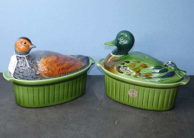 Two ceramic dishes with lids shaped like ducks, one with a brown and gray duck and the other with a green and brown duck, both on a gray surface against a blue background.